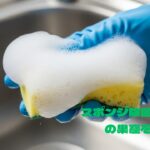 青いゴム手袋をした手が洗剤で泡立てられた黄色と緑のスポンジを握っている様子。背景はステンレス製のシンクの一部。